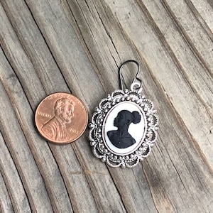 May include: Silver earring with a black silhouette of a woman's profile in a white cameo setting. The cameo is surrounded by a silver filigree frame.