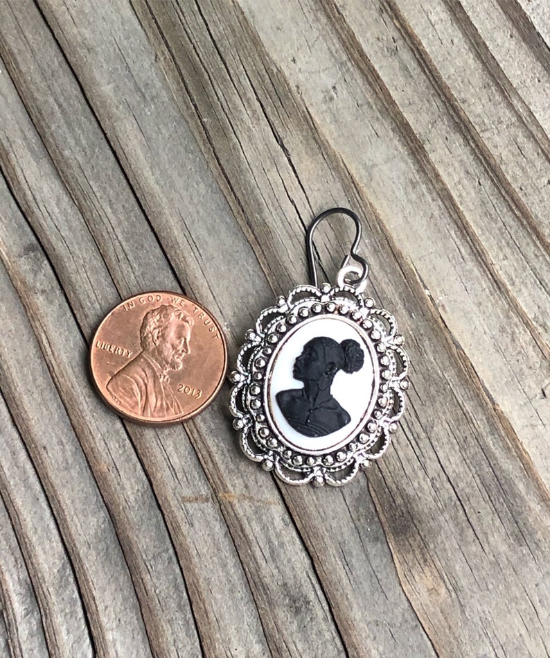 May include: Silver earring with a black silhouette of a woman's profile in a white cameo setting. The cameo is surrounded by a silver filigree frame.