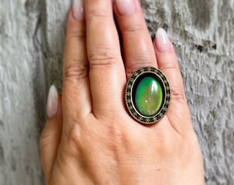Puede incluir: Un anillo ovalado con una piedra que cambia de color. El anillo tiene un marco de color bronce oscuro con un borde detallado. La piedra cambia de verde a amarillo. El anillo se lleva en un dedo.
