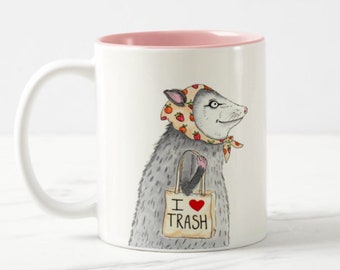 Peut inclure: Une tasse en céramique blanche avec un bord rose. La tasse présente une illustration de dessin animé d'un opossum portant un bandana floral rose et blanc et portant un sac brun qui dit "I ♥ TRASH".