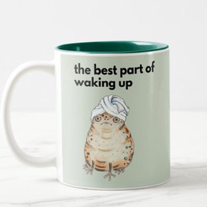 May include: White ceramic mug with a green interior rim and handle. The mug features an illustration of a grumpy-looking frog wearing a towel on its head. The text "the best part of waking up" is printed above the frog.