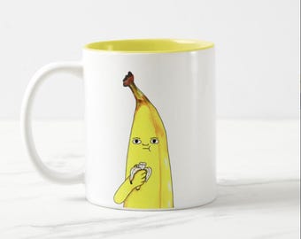 Peut inclure: Mug en céramique blanche avec un intérieur jaune. Le mug présente un personnage de banane de dessin animé avec une expression surprise, tenant une banane pelée. Le personnage de la banane est jaune avec des détails bruns.