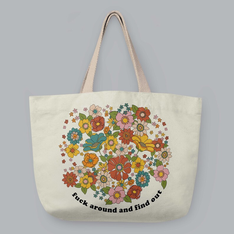 Może przedstawiać: Kremowa torba typu tote z kwiatowym wzorem w stylu retro w odcieniach pomarańczu, ż&oacute;łci, r&oacute;żu i błękitu. Torba ma jasnobeżowe uchwyty i czarny napis "fuck around and find out" wygięty wzdłuż dolnej części kwiatowego wzoru.