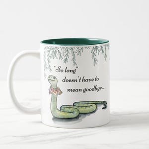 Könnte beinhalten: Weißer Keramikbecher mit grünem Innenraum. Er zeigt eine Illustration einer grünen Schlange mit Blumenkranz und dem Text "So long doesn't have to mean goodbye..." mit Blättern.