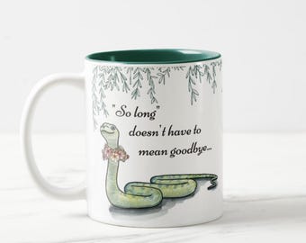 Peut inclure: Mug en céramique blanche avec intérieur et anse verts. L'illustration fantaisiste représente un serpent vert portant une couronne florale, avec le texte "So long doesn't have to mean goodbye..."
