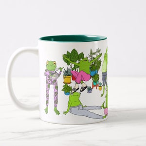 May include: White ceramic mug with a green rim. The mug features a colorful illustration of frogs in various poses, surrounded by plants. The frogs are wearing clothes and some are holding objects.