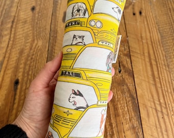 Peut inclure: Un jouet pour chat rectangulaire avec un motif de taxi jaune. Le tissu présente des illustrations de divers chiens et chats dans les taxis. Le jouet est tenu dans une main et se trouve sur un fond en bois.