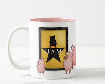 Peut inclure: Mug en céramique blanche avec un bord intérieur rose. Le mug présente un graphisme jaune avec une silhouette de cochon noir et le mot "HAM" en dessous. Trois cochons de dessin animé sont dessinés sur le côté droit du mug, dont un tenant un billet.
