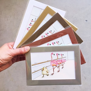 May include: A collection of framed art prints featuring a whimsical illustration of pink flamingos and a small bird in a ballet studio. The frames are in various colors, including white, gold, and brown.