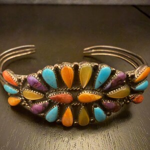 May include: A silver cuff bracelet featuring a cluster of teardrop-shaped stones in shades of turquoise, orange, yellow, and purple. The metal band consists of three parallel wires that meet at the ends, with decorative twisted wire accents.