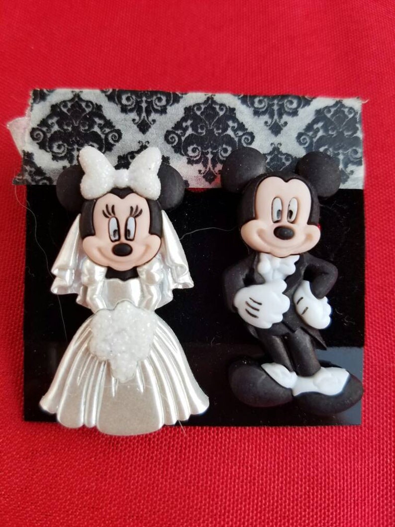 Puede incluir: Un par de figuras de Mickey Mouse y Minnie Mouse con tem&aacute;tica de boda. Mickey lleva un esmoquin y Minnie lleva un vestido de novia blanco con velo y lazo.