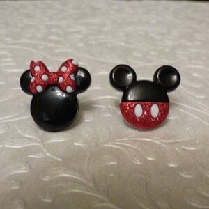May include: A pair of black and red cartoon character earrings. One earring features Mickey Mouse with red glitter and white eyes. The other earring features Minnie Mouse with a red bow and white polka dots.