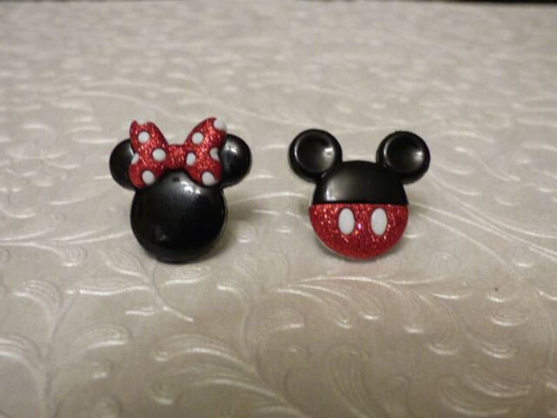 May include: A pair of black and red cartoon character earrings. One earring features Mickey Mouse with red glitter and white eyes. The other earring features Minnie Mouse with a red bow and white polka dots.