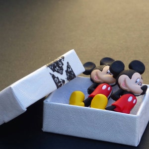 Może przedstawiać: Dwie małe figurki Myszki Miki w białym pudełku z czarno-białym wzorem. Figurki są czerwone, czarne i ż&oacute;łte.