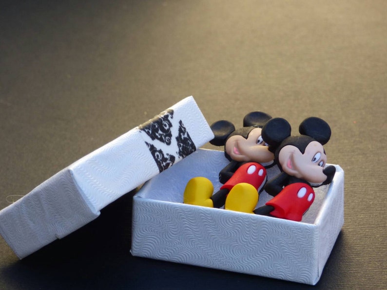 Może przedstawiać: Dwie małe figurki Myszki Miki w białym pudełku z czarno-białym wzorem. Figurki są czerwone, czarne i ż&oacute;łte.