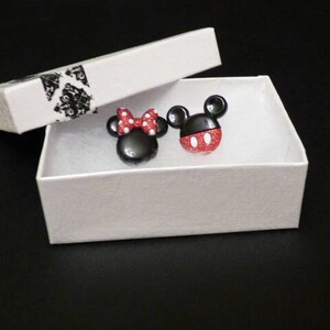 May include: A white box with a black and white patterned lid, containing two small black and red character earrings. The earrings are shaped like Mickey Mouse and Minnie Mouse.