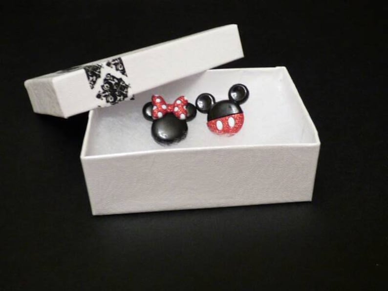 May include: A white box with a black and white patterned lid, containing two small black and red character earrings. The earrings are shaped like Mickey Mouse and Minnie Mouse.