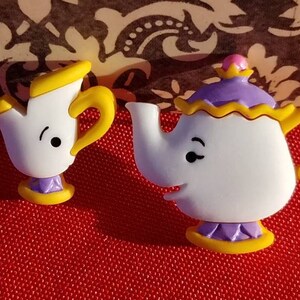 May include: Two ceramic teacups with faces, one yellow and one purple, with a yellow handle. The teacups are from the Disney film Beauty and the Beast.