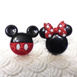 May include: A pair of black and red glitter Mickey Mouse and Minnie Mouse character buttons. Mickey Mouse has black ears and a red and white polka dot shirt. Minnie Mouse has black ears and a red and white polka dot bow.
