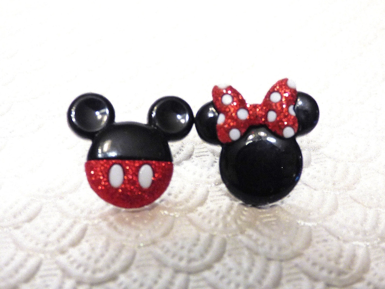May include: A pair of black and red glitter Mickey Mouse and Minnie Mouse character buttons. Mickey Mouse has black ears and a red and white polka dot shirt. Minnie Mouse has black ears and a red and white polka dot bow.