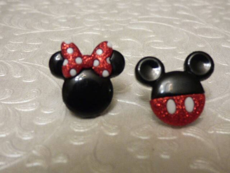 May include: A pair of black and red glitter Mickey Mouse and Minnie Mouse character buttons. Minnie Mouse has a red bow with white polka dots.