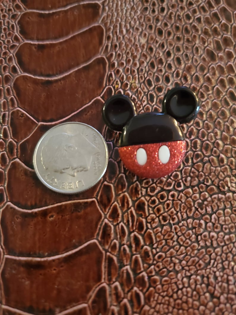 May include: A small, red, black, and white Mickey Mouse figurine with glitter on the red portion. The figurine is about the size of a US quarter.