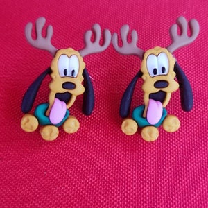 Puede incluir: Dos adornos con forma de perro de dibujos animados con cornamentas, cuerpos amarillos, orejas negras y lenguas rosas. Los perros tienen collares verdes y están sobre un fondo rojo. Estos adornos son probablemente para decoraciones navideñas.