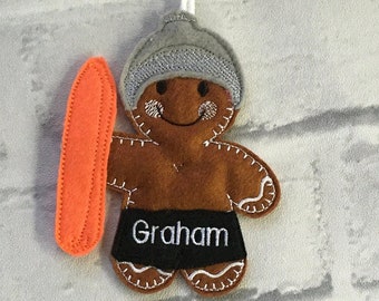 May include: A brown felt gingerbread man ornament wearing a gray hat and black swim trunks with the name "Graham" embroidered on them. The ornament is holding an orange surfboard and has a white string for hanging.