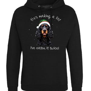 Puede incluir: Sudadera con capucha negra con un perro con un gorro de Navidad. El perro tiene pelaje negro y ojos marrones. El texto "He's making a list" y "I've eaten it twice!" están escritos encima y debajo del perro.