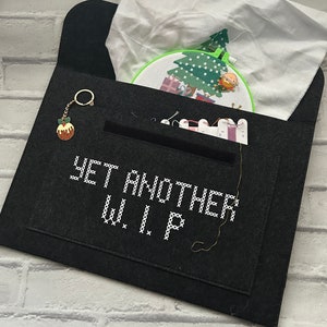 Puede incluir: Bolsa de proyecto de fieltro negro con un diseño bordado en blanco que dice "Yet Another W.I.P." La bolsa tiene un bolsillo y una cremallera. Un pequeño aro de bordado de un árbol de Navidad está dentro de la bolsa.