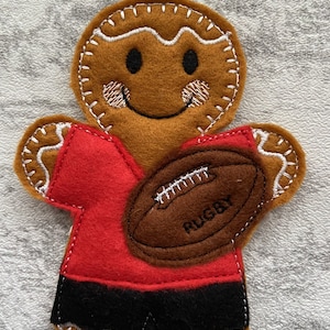 May include: A gingerbread man wearing a red jersey and black shorts, holding a brown rugby ball with the word "RUGBY" embroidered on it.