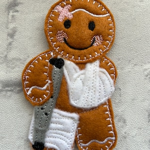 May include: A brown gingerbread man ornament with a white bandage on his head, a white cast on his arm, and a white cast on his leg. He is holding a grey crutch.