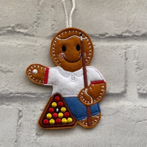 May include: A gingerbread person ornament with a brown felt body, white felt shirt, blue felt trousers, and a brown felt cue stick. The gingerbread person is holding a brown felt triangle with red, yellow, and black felt balls representing a snooker rack.