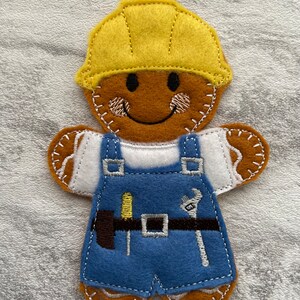 May include: A gingerbread person wearing a yellow hard hat, white shirt, blue overalls, and a tool belt with a hammer, screwdriver, and wrench.