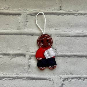 May include: A gingerbread person ornament with a red shirt, blue trousers, and a white gift in its hand. The ornament is brown and has a white string for hanging.