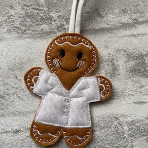 Peut inclure: Une décoration de pain d'épices en forme de personnage avec un manteau blanc et un visage souriant. La décoration est en feutre et possède une ficelle blanche pour l'accrocher.