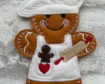 May include: A gingerbread man ornament with a white chef's hat, apron, and rolling pin. He is holding a gingerbread man in his apron pocket and has a red heart on his apron.
