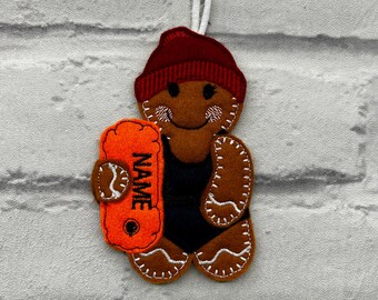 May include: A felt gingerbread man ornament wearing a maroon beanie and black swimsuit, holding an orange rescue buoy. The buoy has the word "NAME" printed on it. The ornament is brown with white stitching and a white string for hanging.