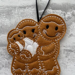 May include: A felt gingerbread family ornament featuring a brown gingerbread man, a brown gingerbread woman, and a white gingerbread baby. The ornament has a black ribbon for hanging.