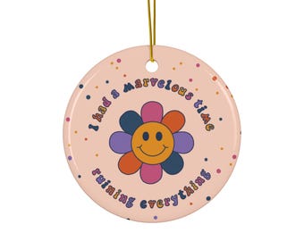 May include: Round ceramic ornament with a peach background and a retro flower design. The flower has orange, pink, purple, and blue petals with a yellow smiley face center. The text "I had a marvelous time ruining everything" is written around the edge.