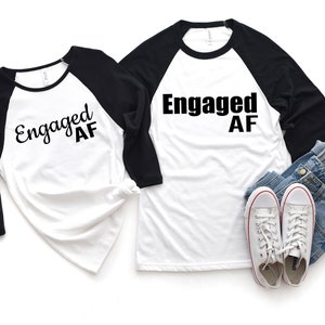May include: Two white baseball t-shirts with black sleeves. The shirts have the text "Engaged AF" printed on them in black. The shirts are laid out on a white background.