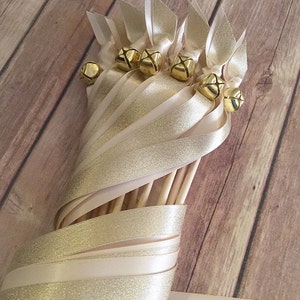 May include: A bundle of ivory and gold ribbon wands with gold bells attached. The wands are tied together with a white ribbon.