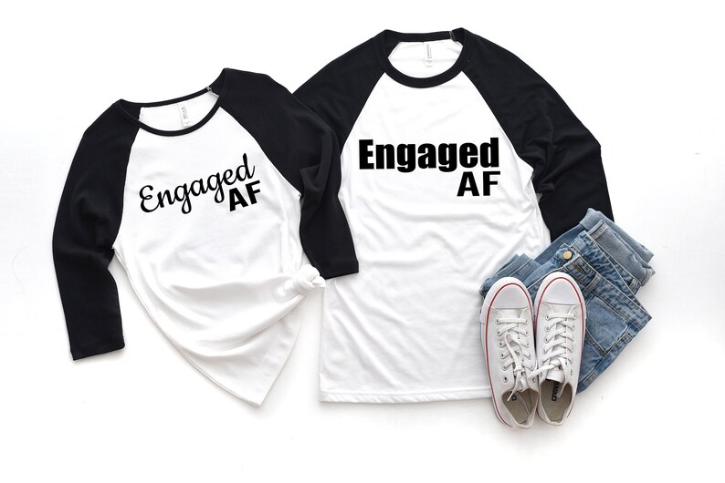 May include: Two white baseball t-shirts with black sleeves. The shirts have the text "Engaged AF" printed on them in black. The shirts are laid out on a white background.