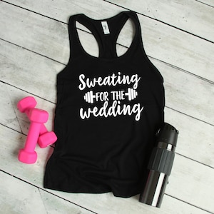 May include: Black tank top with white text that reads "Sweating for the wedding" with a dumbbell graphic.  Two pink dumbbells and a silver water bottle are on a white wood background.