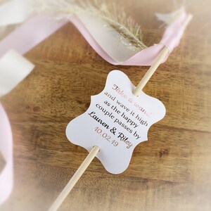 May include: White card with a pink and gold design, attached to a wooden stick. The card has the text "Take a wand and wave it high as the happy couple passes by Lauren & Riley 10.02.19".