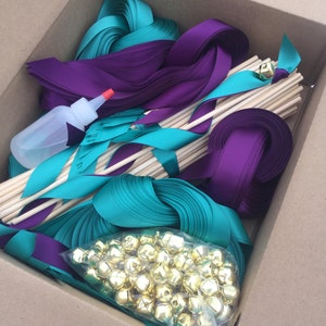 May include: A cardboard box filled with teal and purple ribbon, wooden dowels, and gold bells. The supplies are for making ribbon wands.