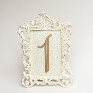 May include: White ornate frame with a gold glitter number one on a white background. The frame is a table number for a wedding or event.