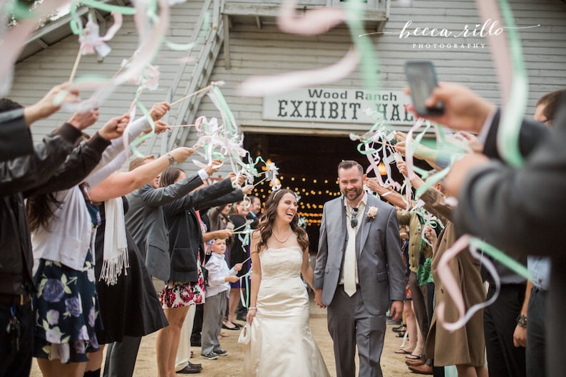 May include: A bride and groom walk down the aisle, surrounded by guests holding colourful ribbons. The sign above the entrance reads "Wood Ranch Exhibit Hall".