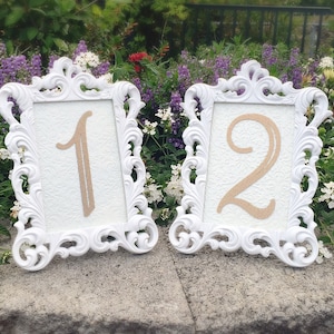 May include: Two white ornate frames with a decorative design, each holding a number. The first frame displays the number "1" and the second frame displays the number "2".