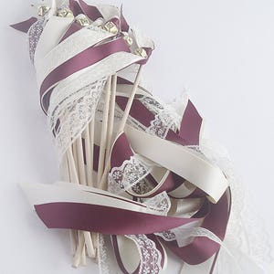 May include: A bundle of burgundy and ivory ribbon wands with lace trim and gold bells. The wands are tied together with a burgundy ribbon and are ready for a wedding or special event.